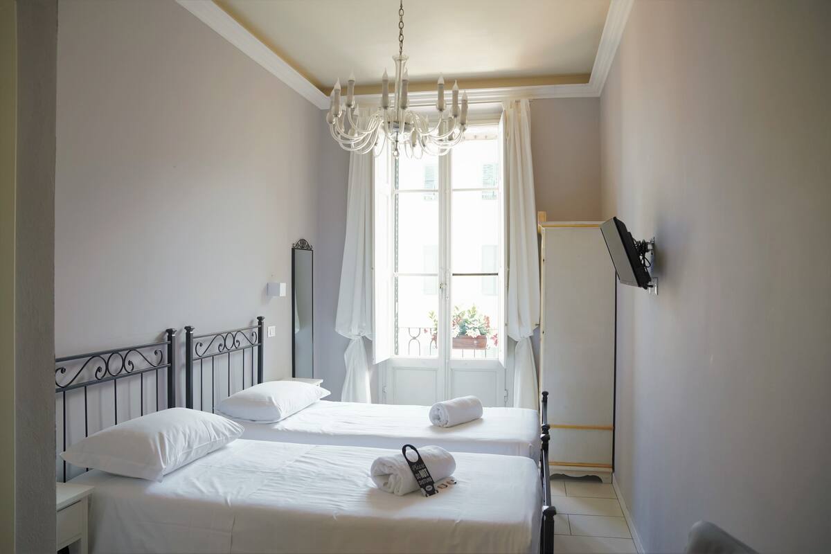 A hotel room with two beds, a chandelier, a large window with curtains, and a flat-screen TV mounted on the wall.