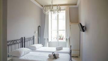Habitación de hotel con dos camas, un candelabro, un ventanal grande con cortinas y un televisor de pantalla plana montado en la pared.