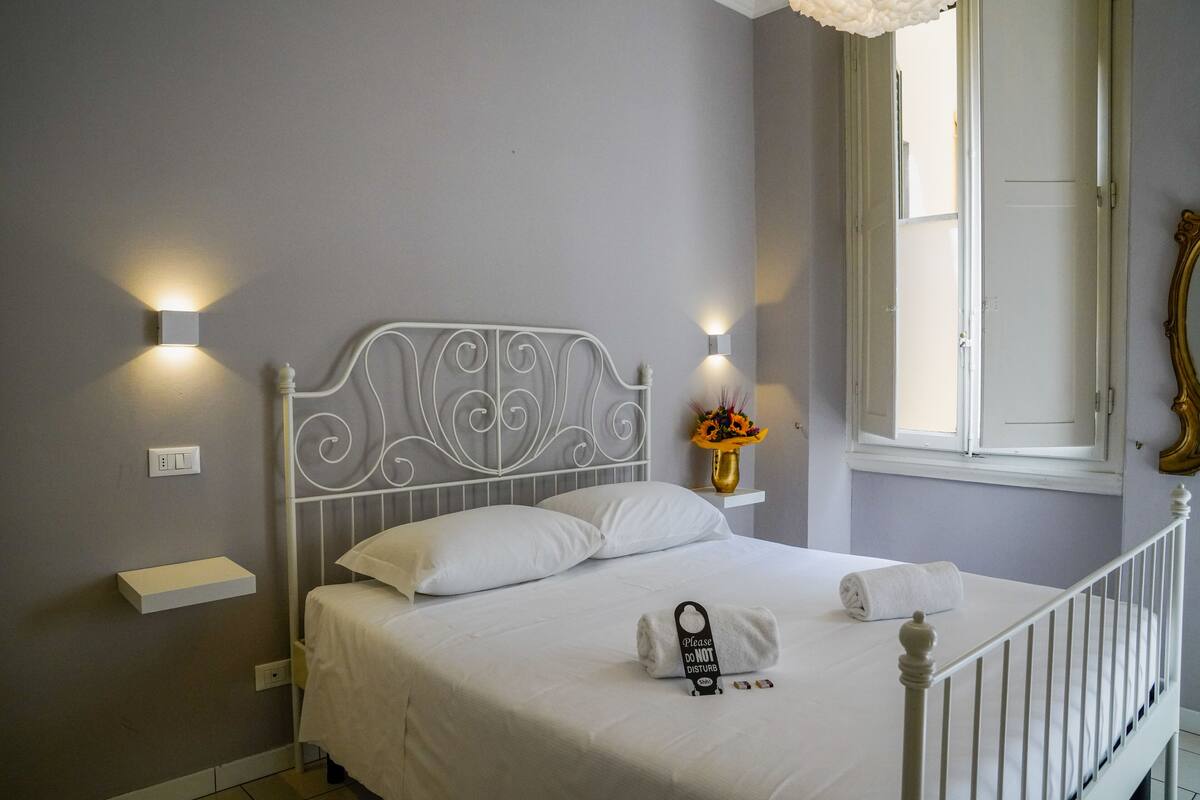 A neatly made bed with white linens, a bedside table with a vase of flowers, and wall-mounted lights.