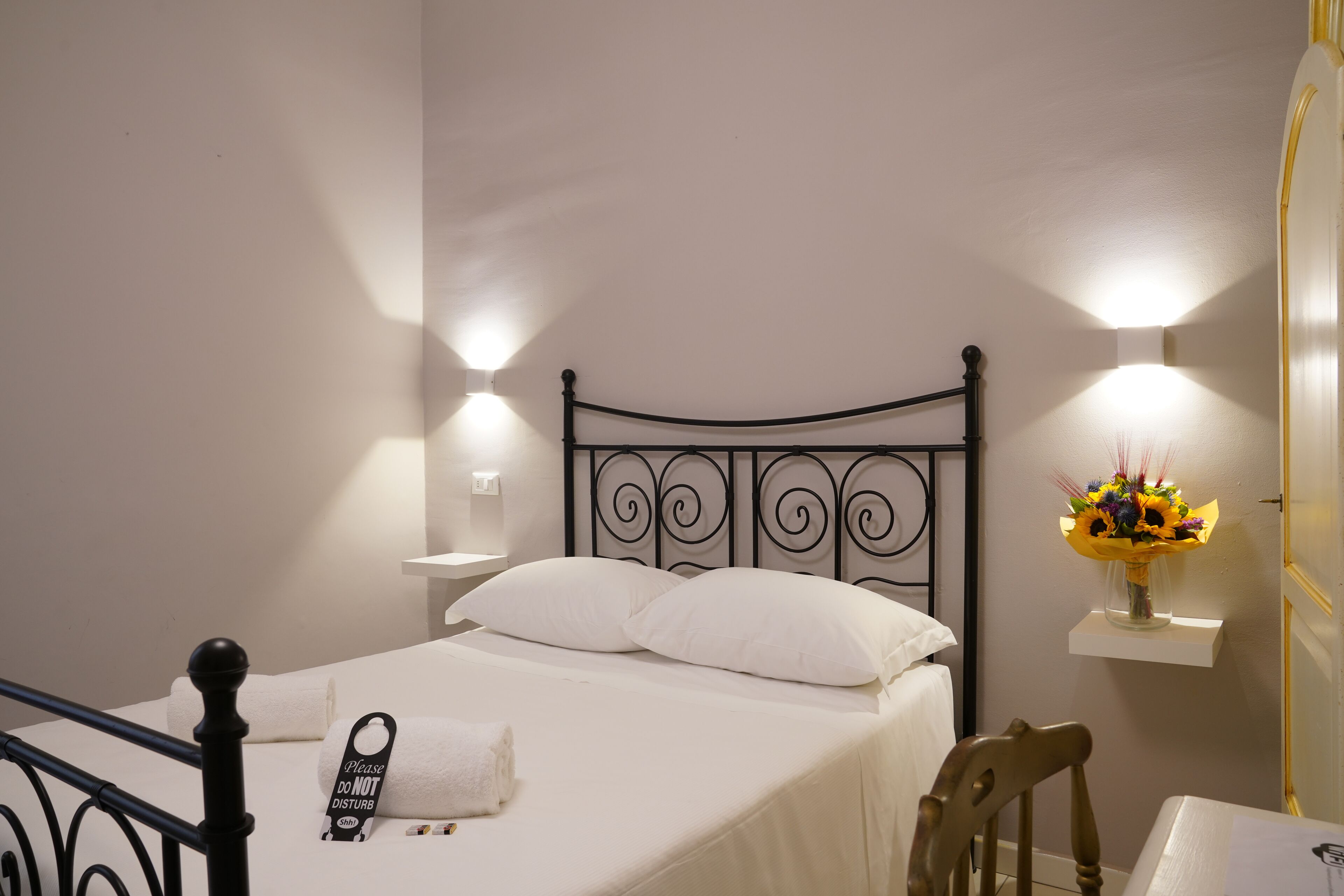 A neatly made bed with white linens and a black metal headboard, a bedside table with a lamp, and a small shelf with a vase of flowers.