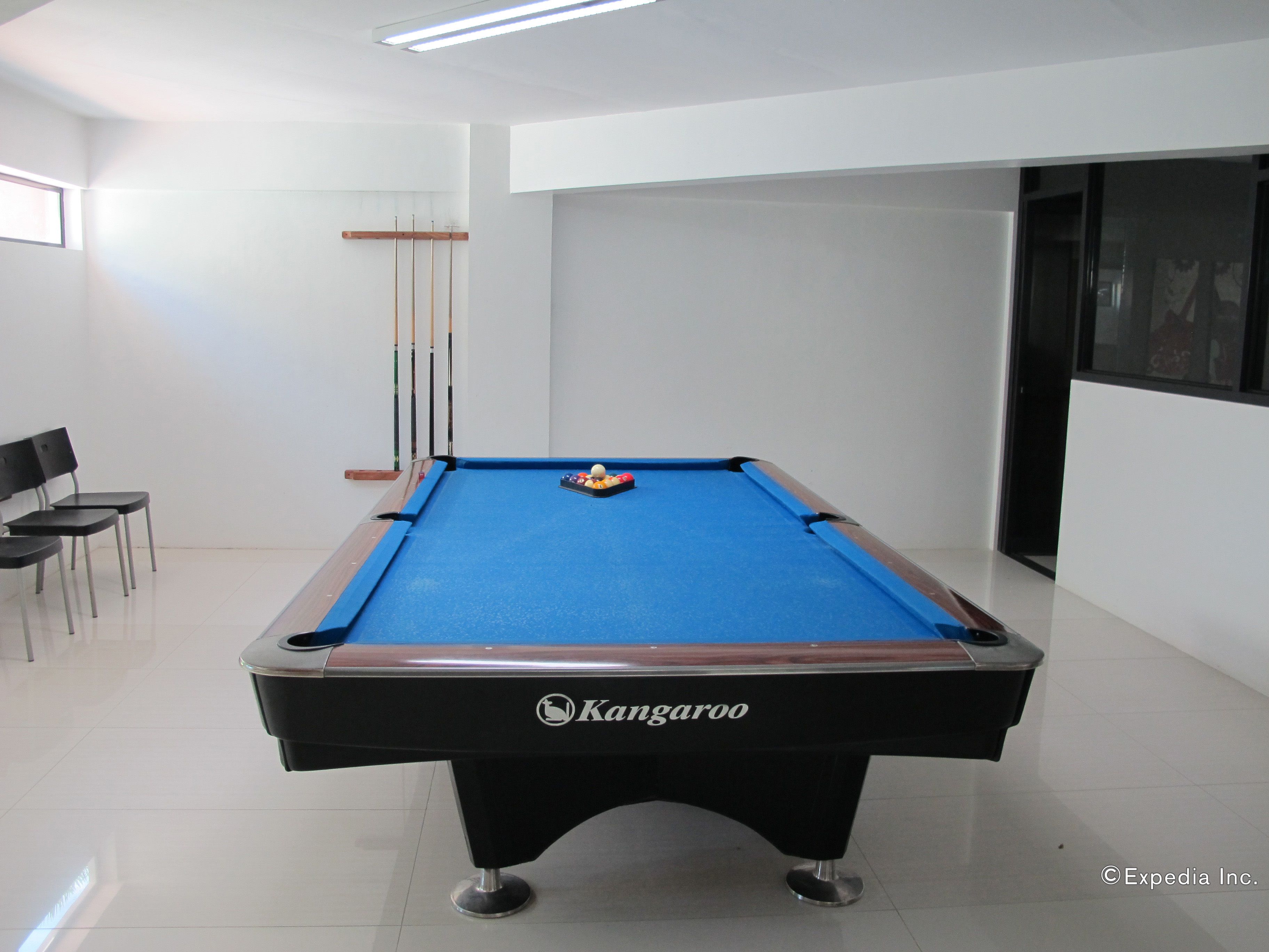 Een pooltafel met een blauwe viltkleed, een Kangaroo logo, en een biljarthouder met biljartkeus. Er staan stoelen en een ingelijst beeld op de muur.