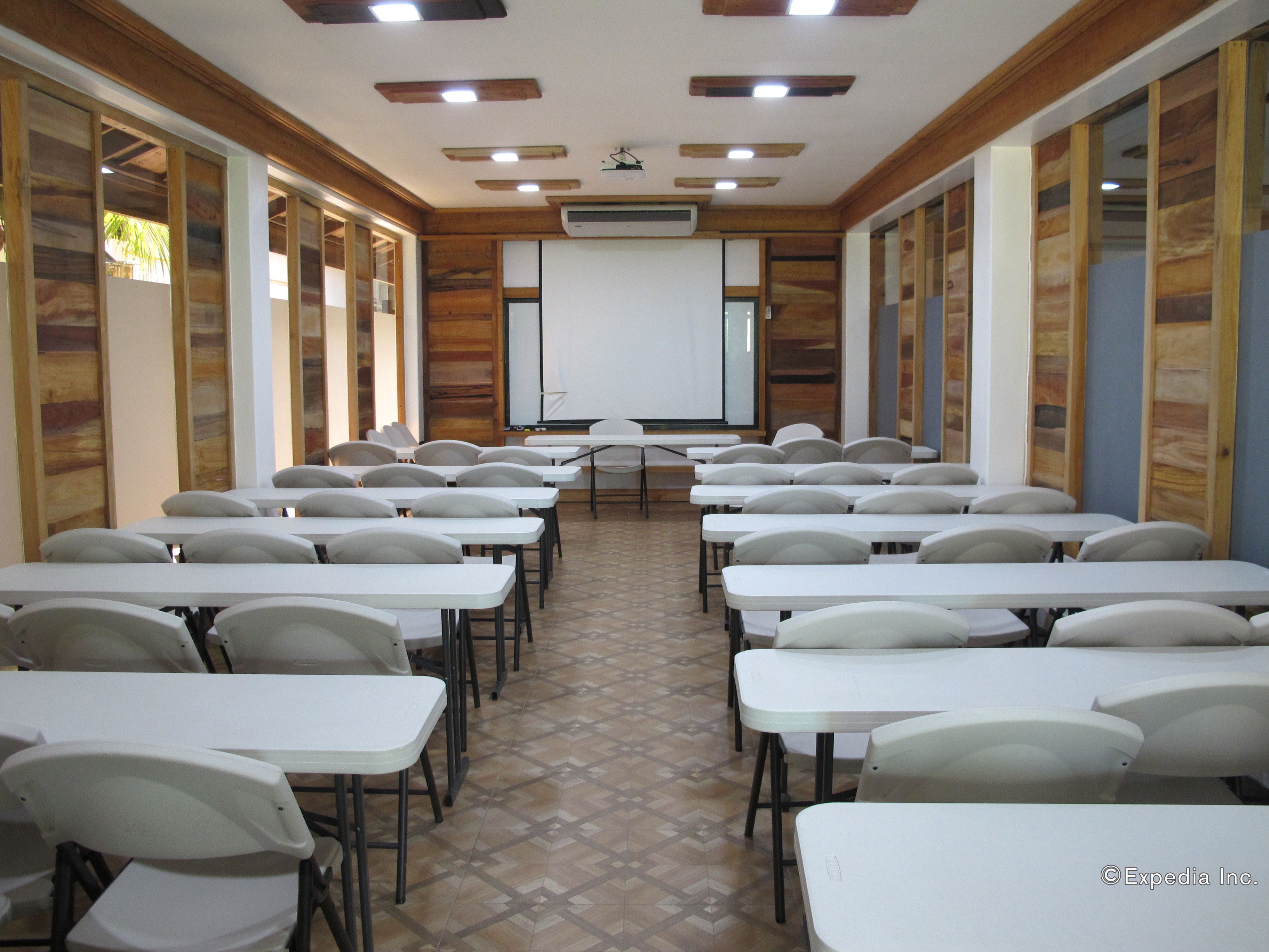 Un aula con mesas y sillas blancas dispuestas en filas, una pantalla de proyector al frente y paneles de madera en las paredes.