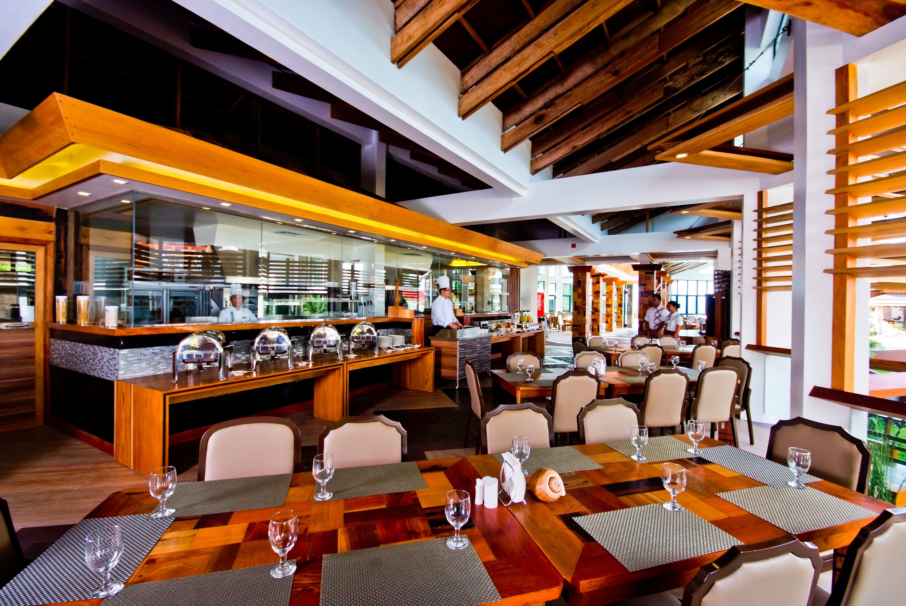 A modern restaurant with wooden tables and chairs, a bar area, and a view of the kitchen.