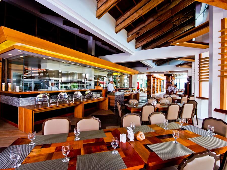 A modern restaurant with wooden tables and chairs, a bar area, and a view of the kitchen.