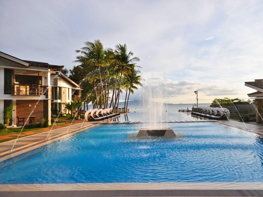 A swimming pool with a central fountain, surrounded by palm trees and lounge chairs, with buildings on either side.