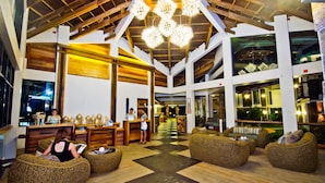 A spacious lobby with high ceilings, wooden beams, and a central chandelier. There are several wicker seating areas and a reception desk with staff.