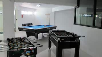A room with a pool table, two foosball tables, and a window with blinds.