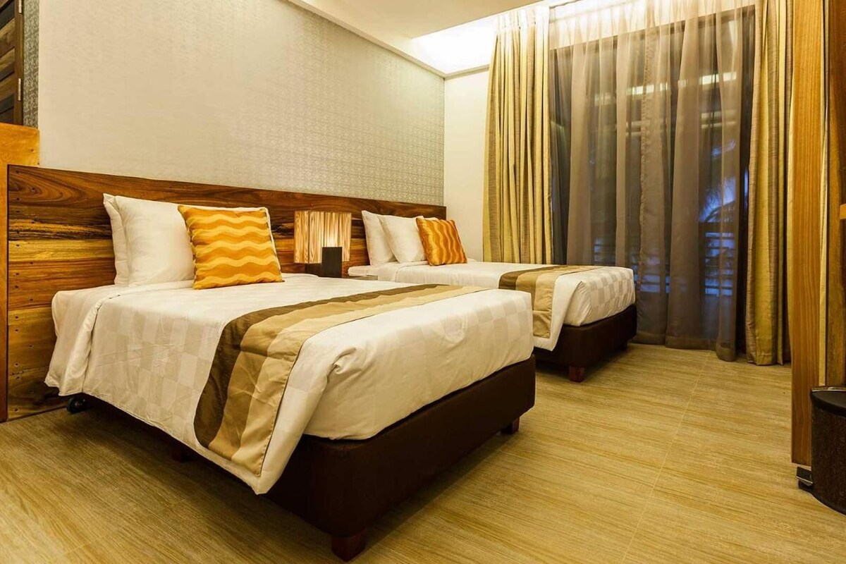 A hotel room with two beds, a wooden headboard, a large window with sheer curtains, and a trash bin.