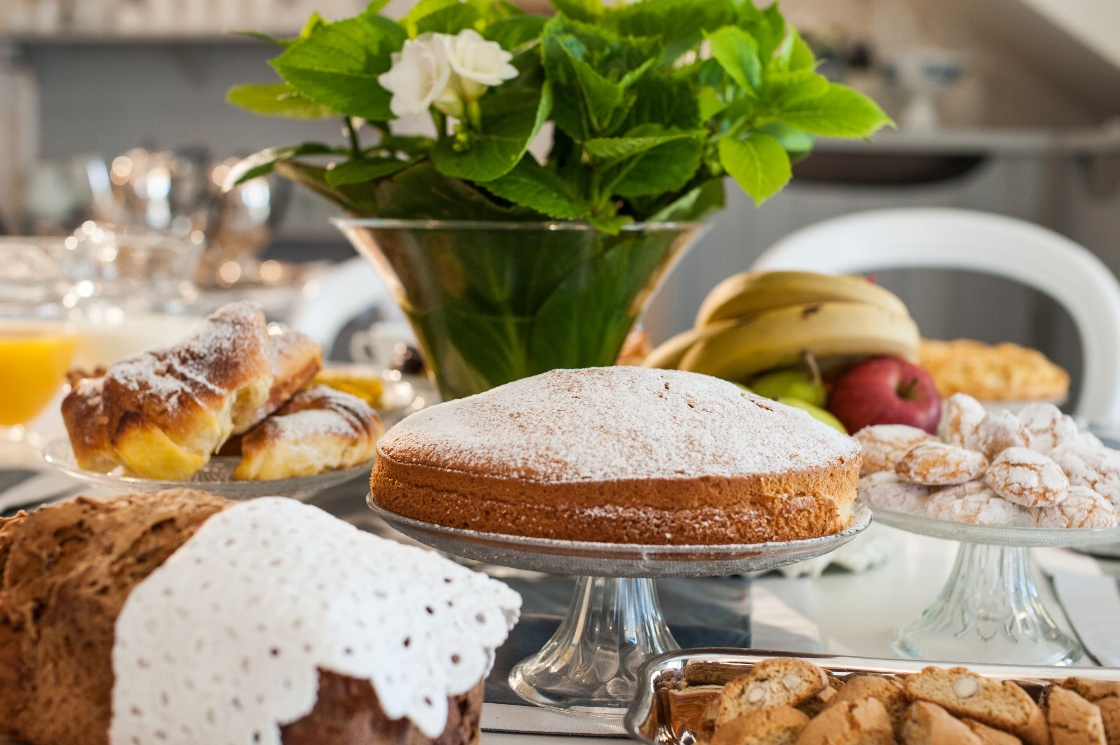 Una mesa con diversos pasteles y un pastel, un cuenco con plantas verdes, y un frutero con plátanos y manzanas.