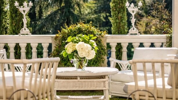 Un área de estar al aire libre cubierta, con muebles de mimbre blanco, una mesa con un jarrón de flores y dos candelabros.