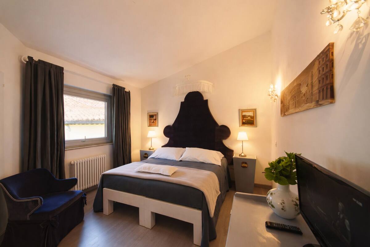 A hotel room with a bed, bedside tables, a chair, a radiator, a window with curtains, a plant, and artwork on the walls.