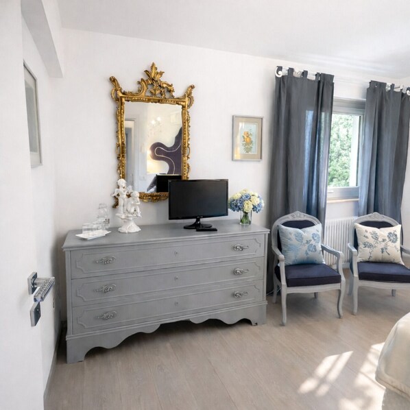 Une pièce comprenant une commode grise, un téléviseur, deux fauteuils avec des coussins à motifs floraux, un grand miroir décoratif et une fenêtre avec des rideaux.
