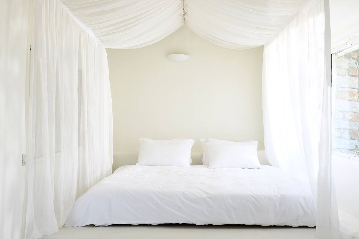 Una cama con dosel blanco, con ropa y almohadas blancas, frente a una pared lisa con un ventanal pequeño.