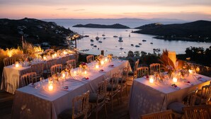 An outdoor dining area with tables set for a meal, overlooking a body of water with boats. - Aigis Suites (Kea)