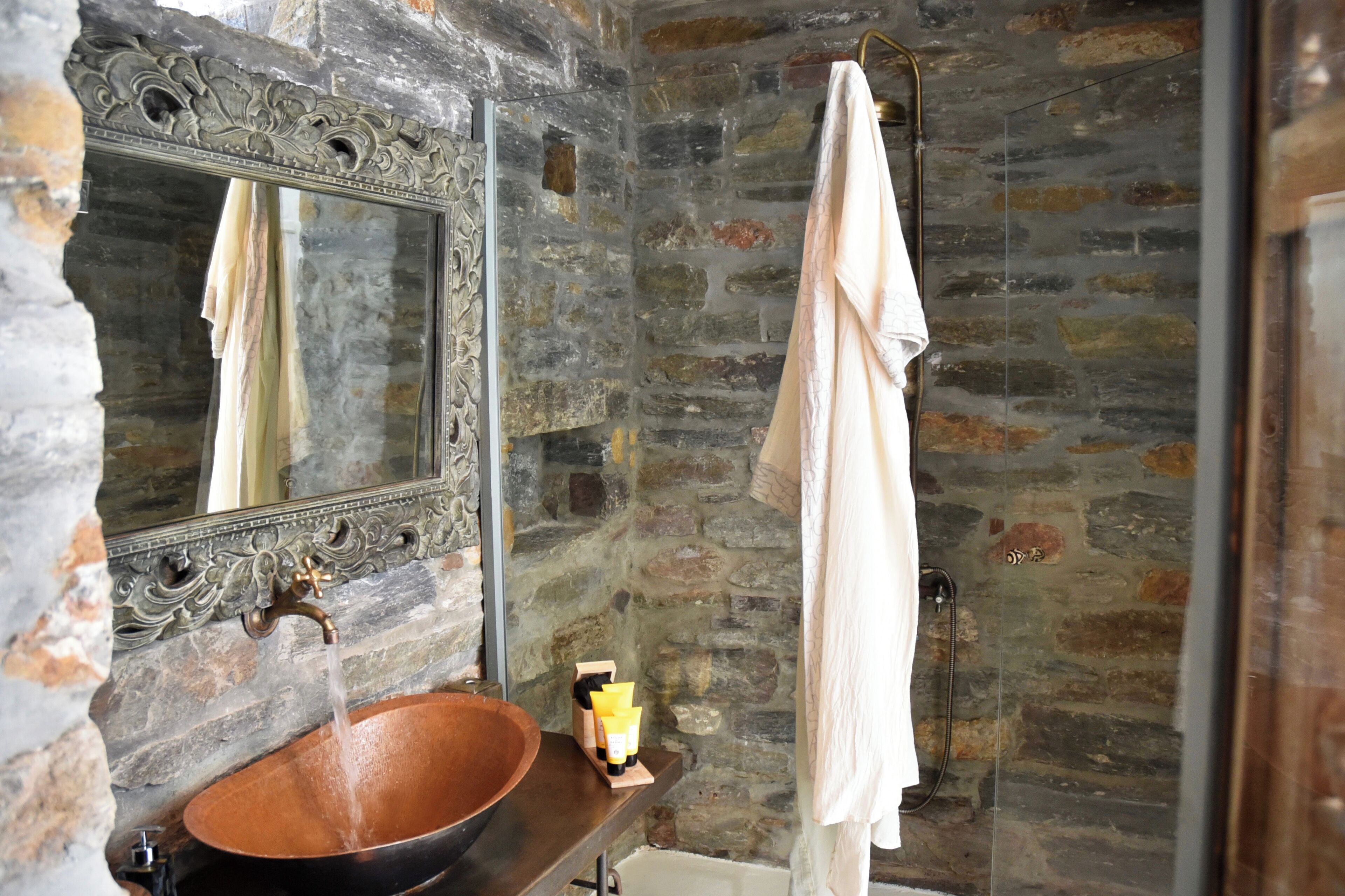 A bathroom with a stone wall, a copper sink, a mirror, and a towel hanging on the wall.
