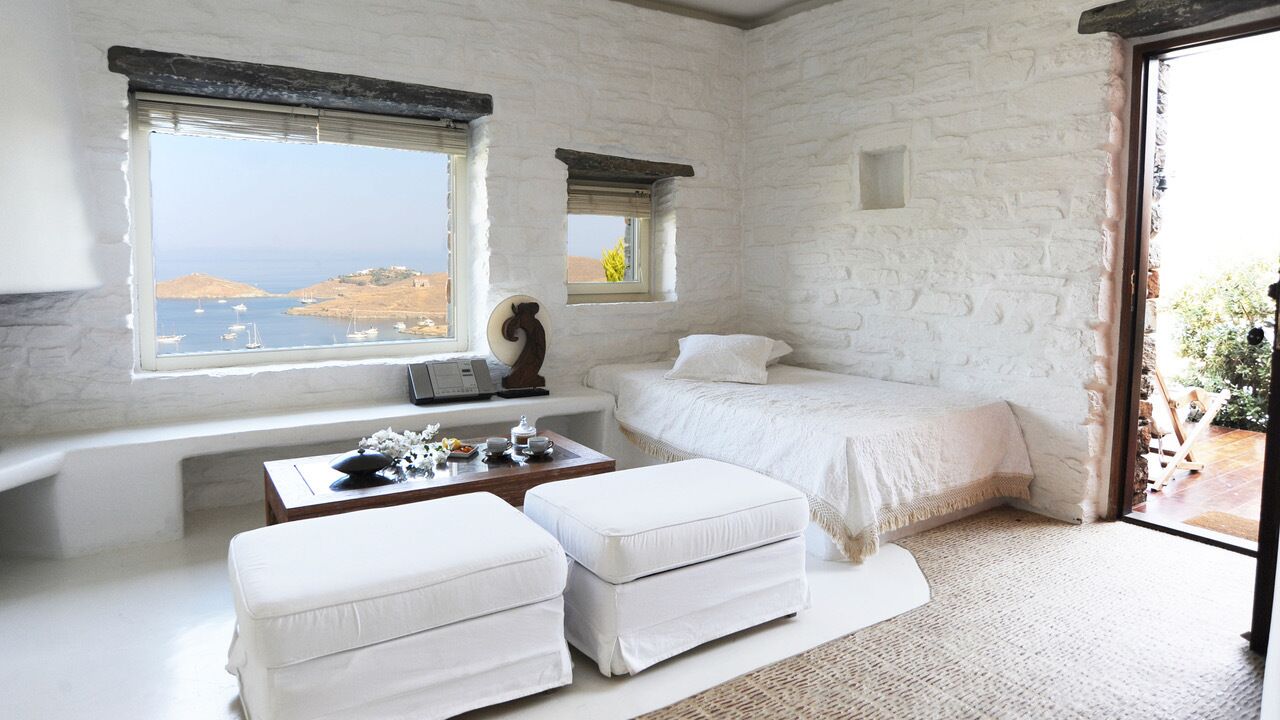 A bedroom with a stone wall, a bed, a desk with a lamp, and a view of the sea.