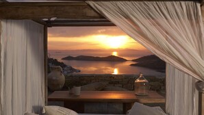 A room with a bed, a table, and a view of a sunset over water. - Aigis Suites (Kea)