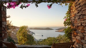 A stone-walled outdoor seating area with two wicker chairs, overlooking a coastal landscape with boats and hills. - Aigis Suites (Kea)