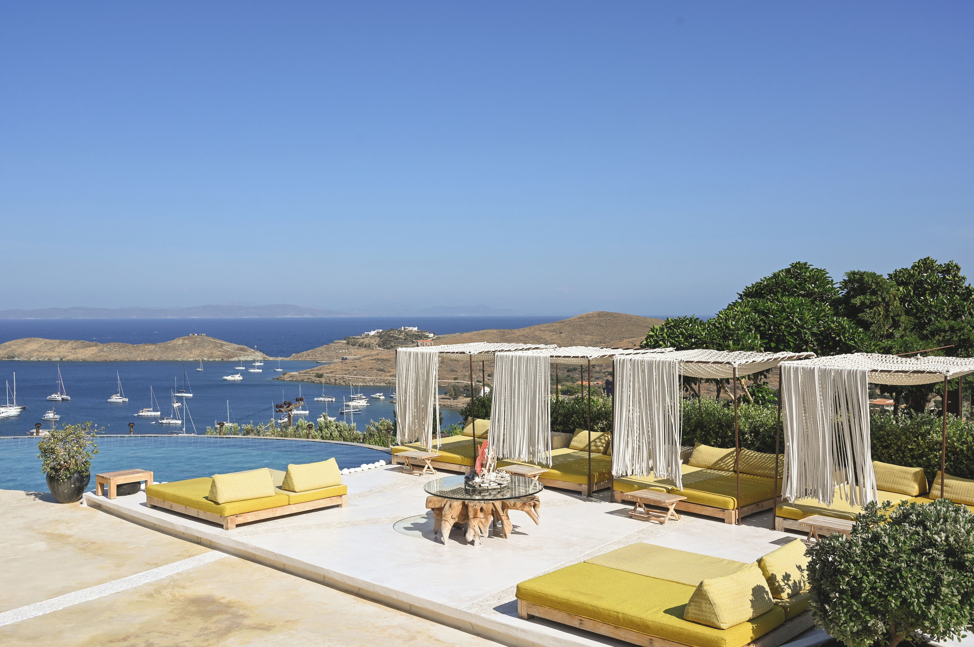 A rooftop terrace with yellow cushioned seating, a round table, and a view of the sea and boats.