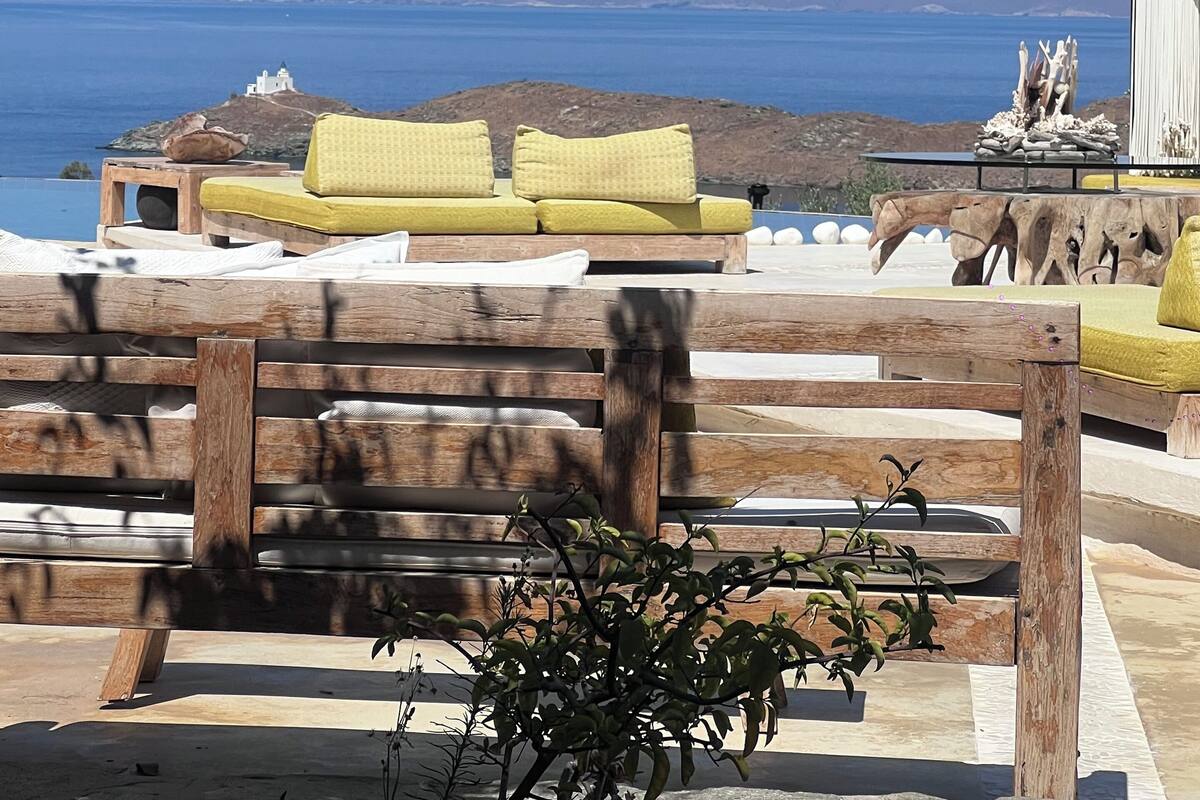 Un banco de madera con cojines, una mesa de madera y vistas al mar y colinas a lo lejos.