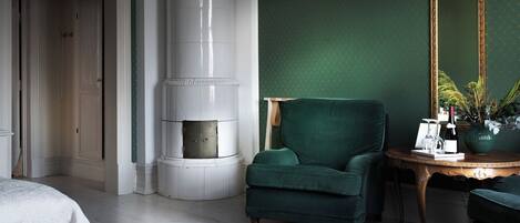 A room with a green wall, a green armchair, a white fireplace, and a wooden side table with a vase and a bottle.