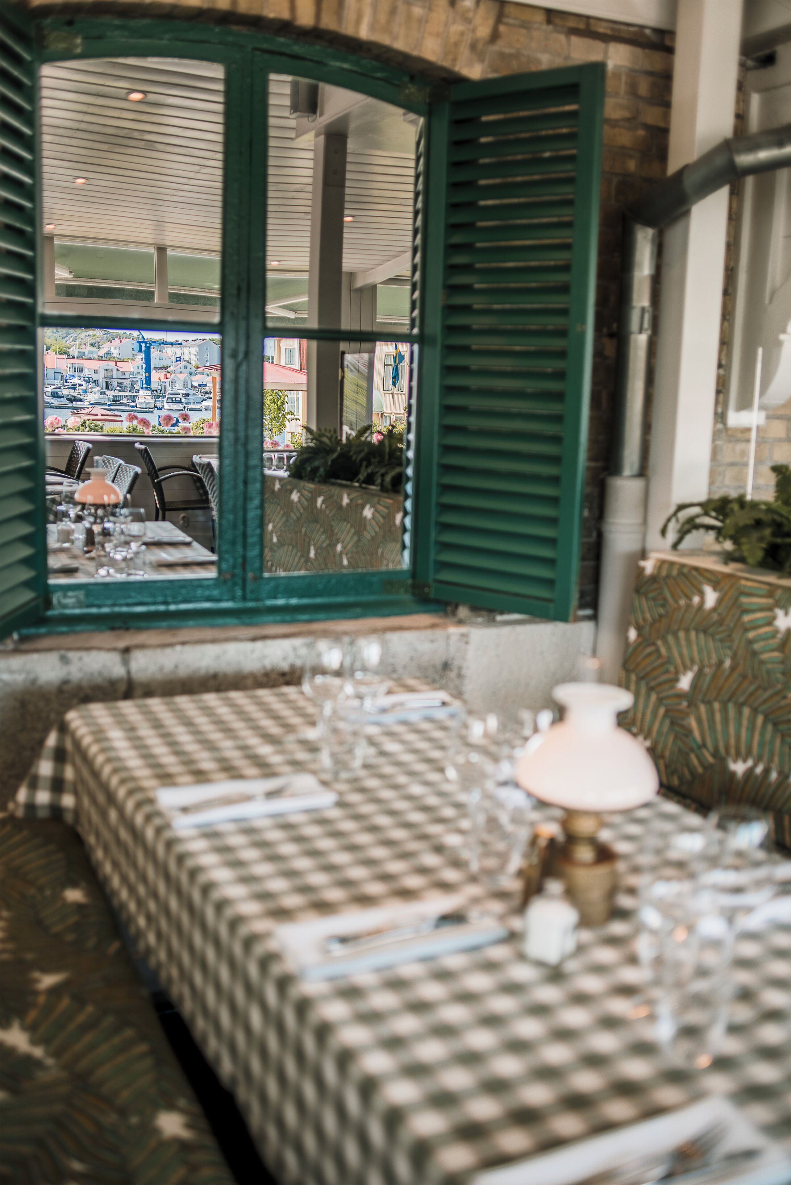 A dining area with a checkered tablecloth, a lamp, and a view through a window with green shutters.