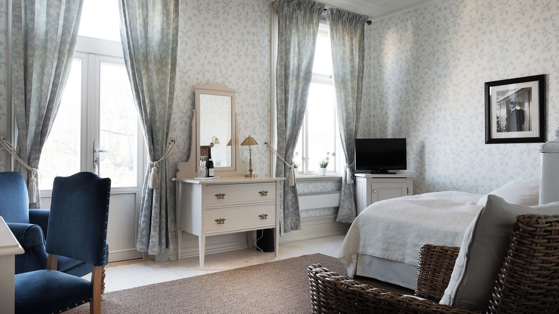 Une chambre à coucher avec un lit, une commode, une chaise, un téléviseur et une fenêtre avec des rideaux.