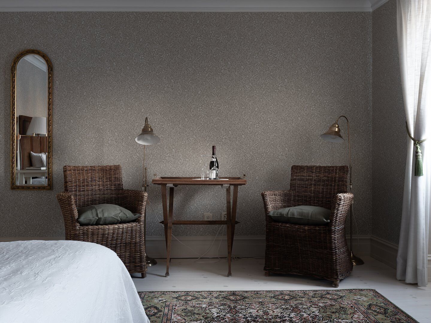 A room with a bed, two wicker chairs, a small table with a bottle, and a mirror on the wall.