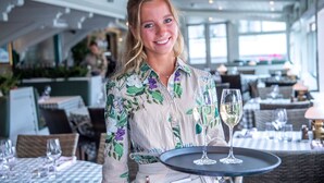 A person holding a tray with two glasses of white wine in a well-lit indoor dining area with tables and chairs.