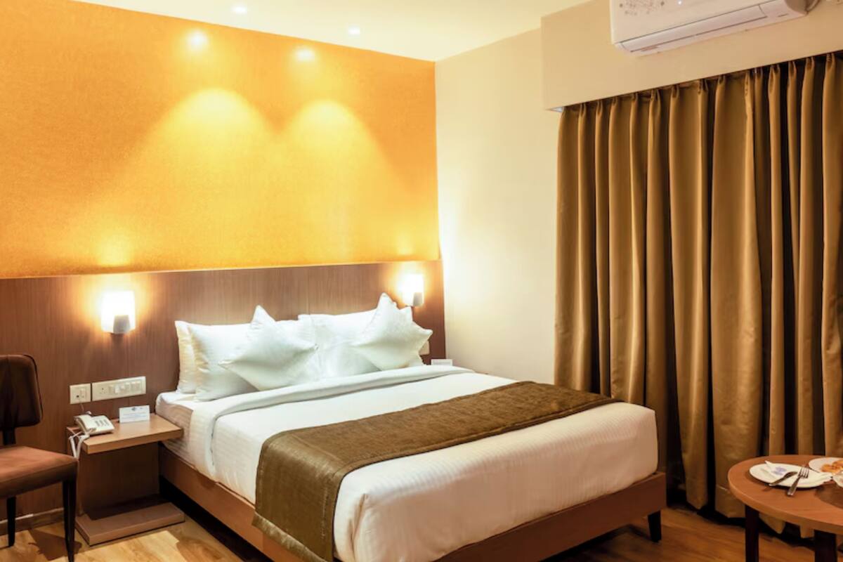 A hotel room with a bed, bedside table, lamp, curtains, and an air conditioning unit.