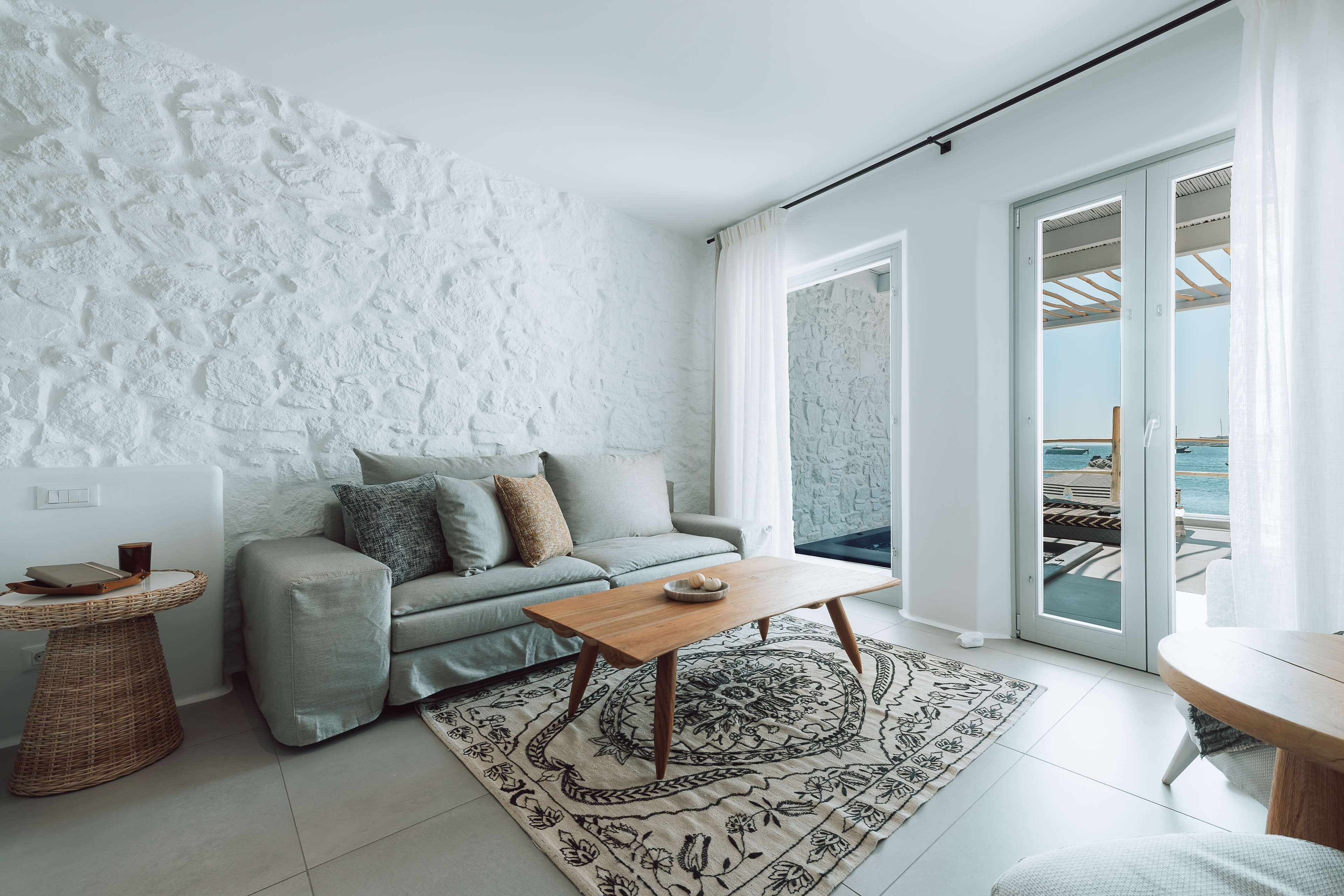 Een moderne woonkamer met een lichtgrijze bank, een houten salontafel, een vloerkleed met een patroon en uitzicht op zee door grote schuifdeuren.
