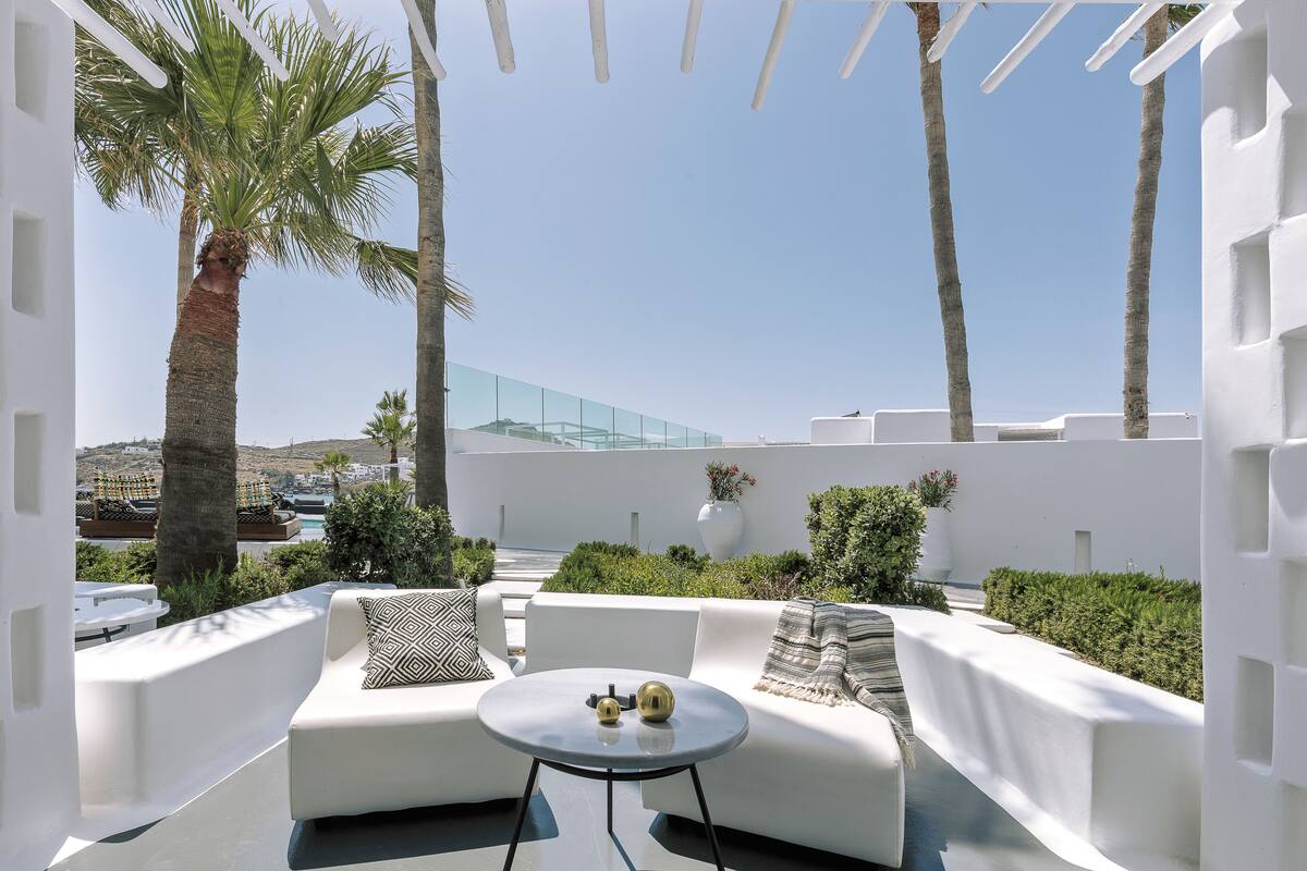 A modern outdoor lounge area with white furniture, a round table, and a view of the sea and buildings.