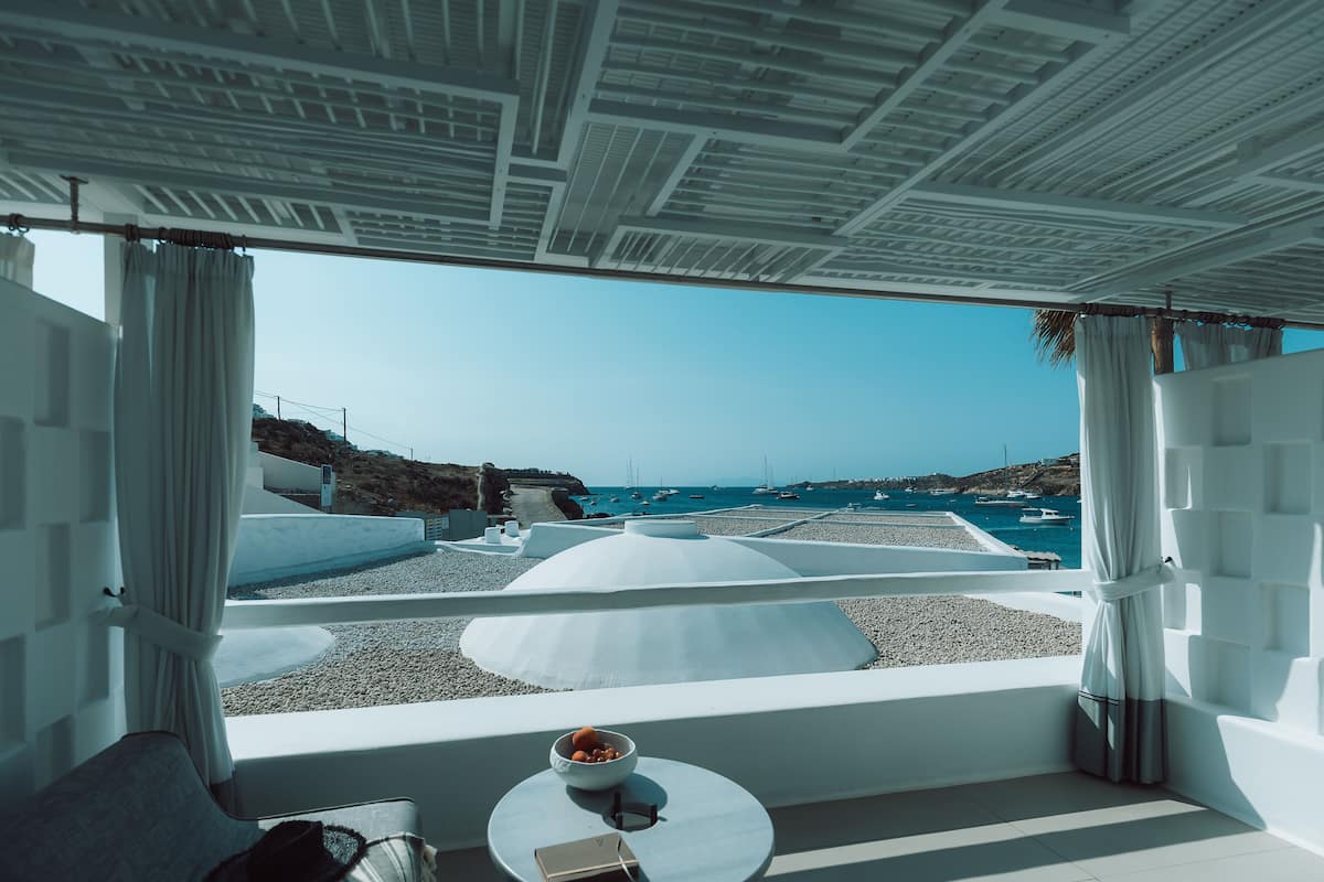 A modern, minimalist room with a view of the sea and boats. It features a white ceiling, white walls, and a round table with a bowl and glasses.