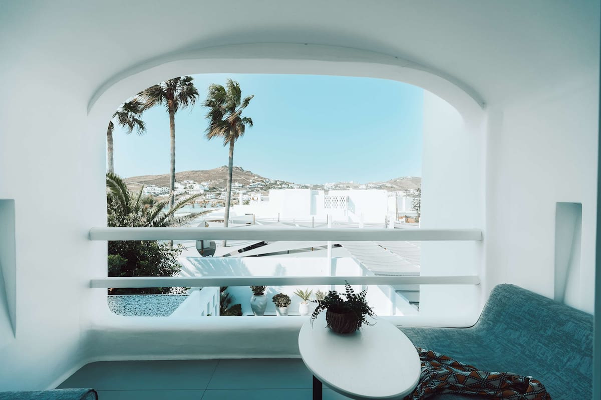 A room with a white balcony overlooking a coastal town, a white table, and a patterned cushion.