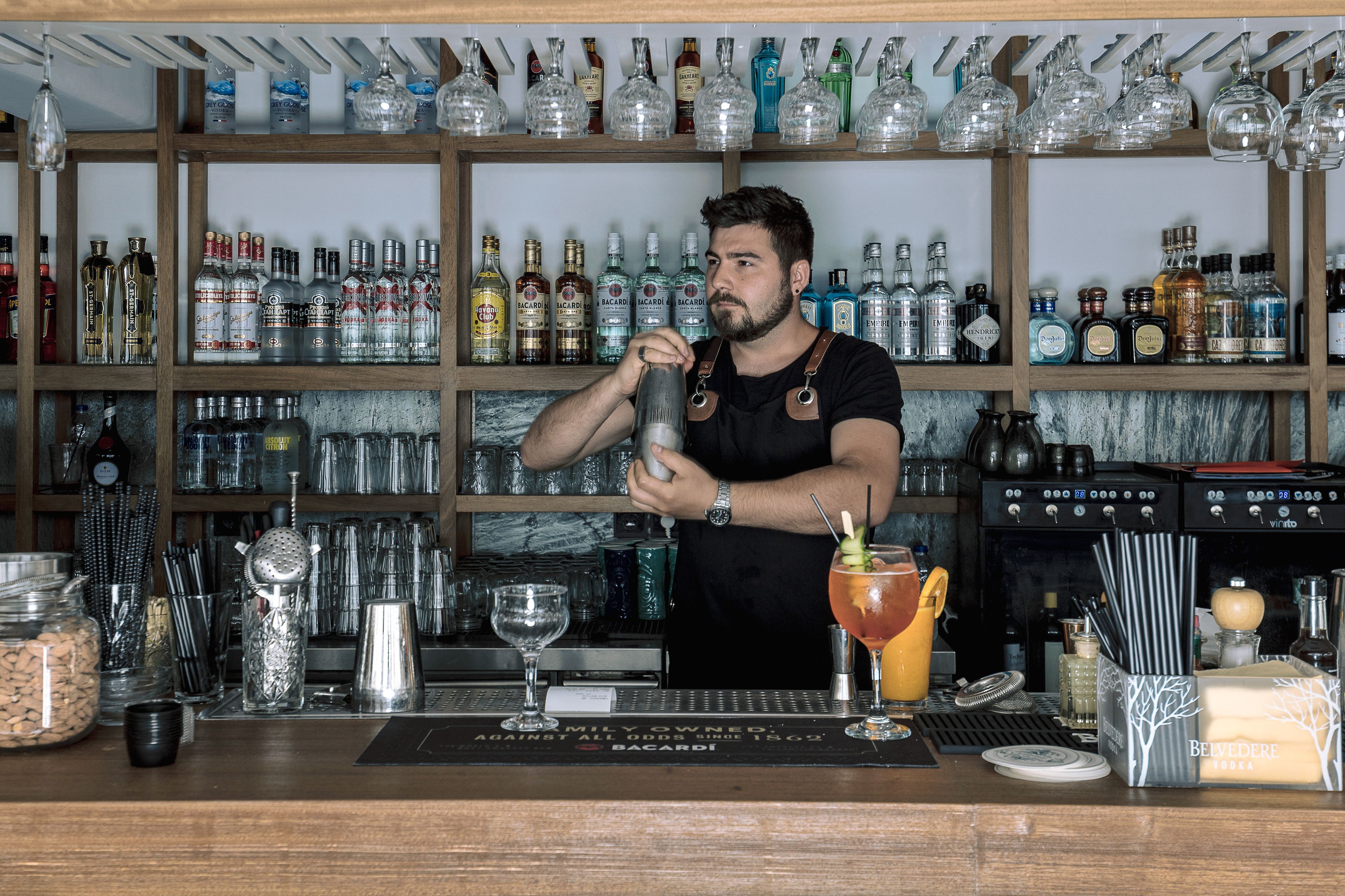 Un cantinero preparando una bebida en la barra, con estantes llenos de botellas de licor y diferentes utensilios.