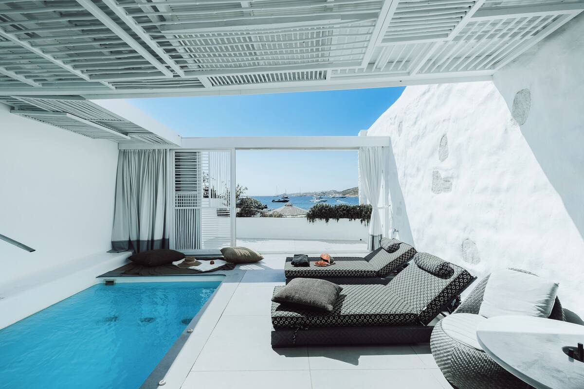 A modern outdoor pool area with a sofa and a table, a clear view of the sea, and a white exterior wall.