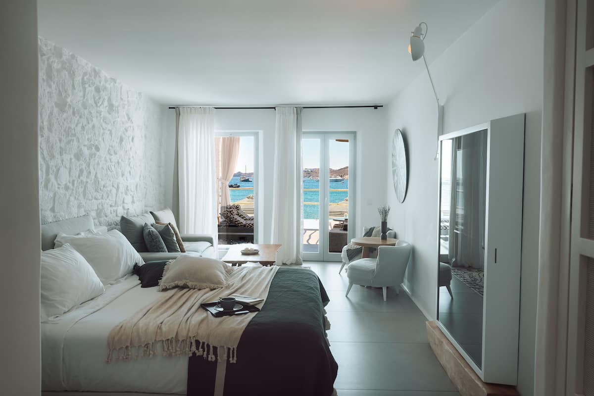 A modern bedroom with a bed, a small table, a chair, and a view of the sea through large windows.
