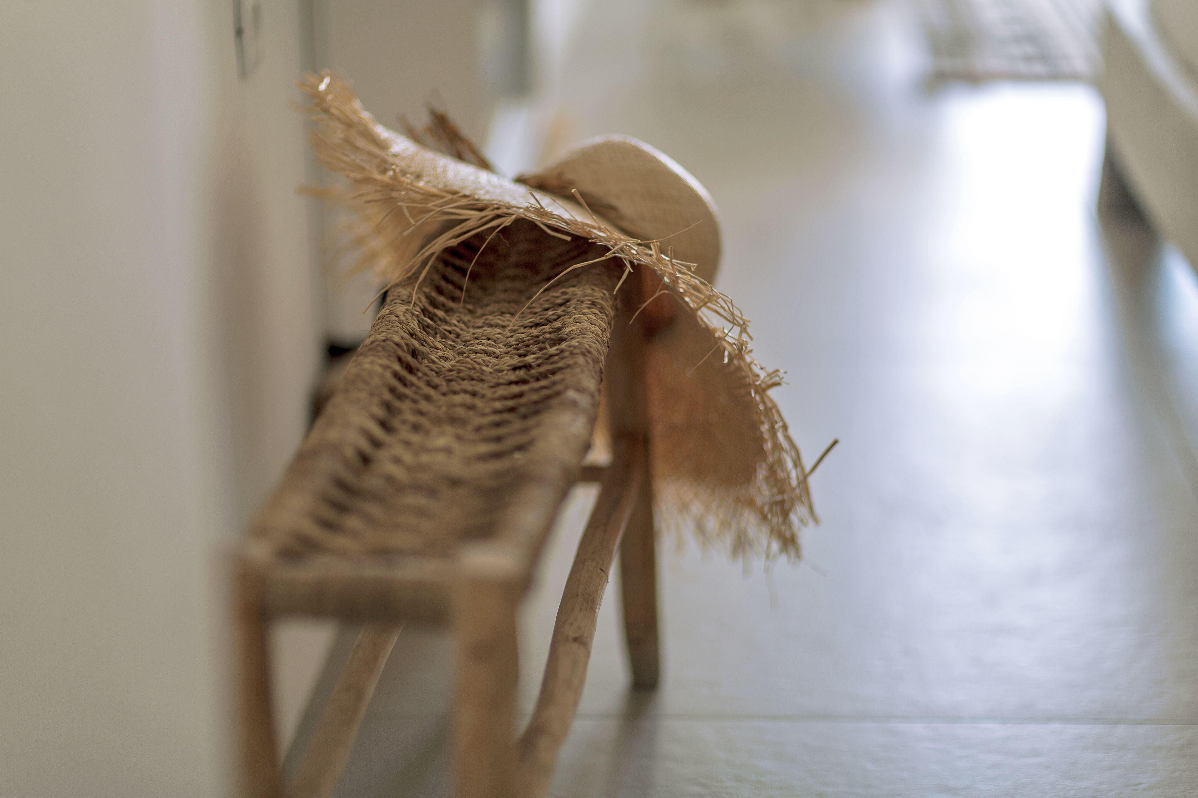 Una silla de mimbre con respaldo trenzado y asiento de madera, ubicada en un pasillo con piso de baldosas.