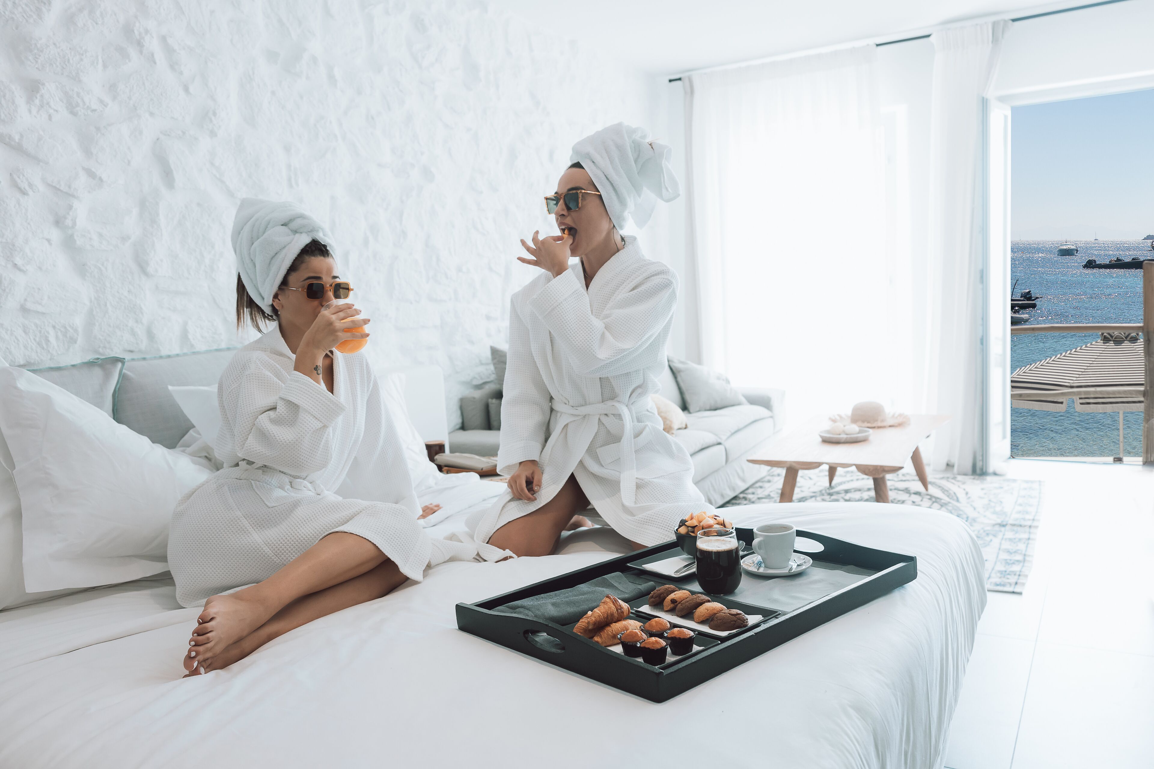 A modern hotel room with a large bed, a breakfast tray with pastries and coffee, and a view of the sea through the window.