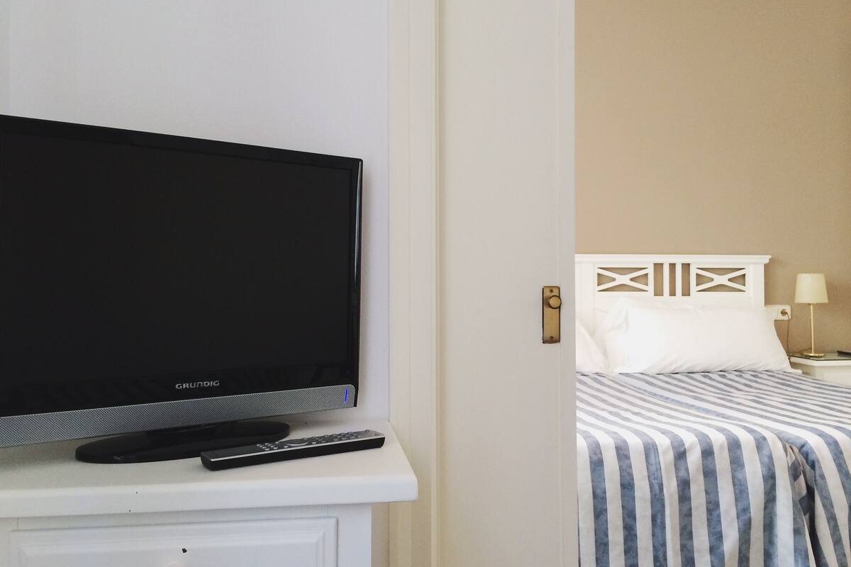 Uma televisão sobre um suporte branco, um controle remoto, uma cama com colcha listrada, uma cabeceira branca e um abajur.