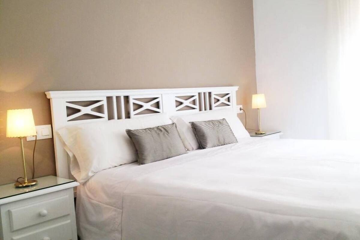 Cama bem arrumada com cabeceira branca, duas luminárias de mesa e mesa de cabeceira branca com gavetas.