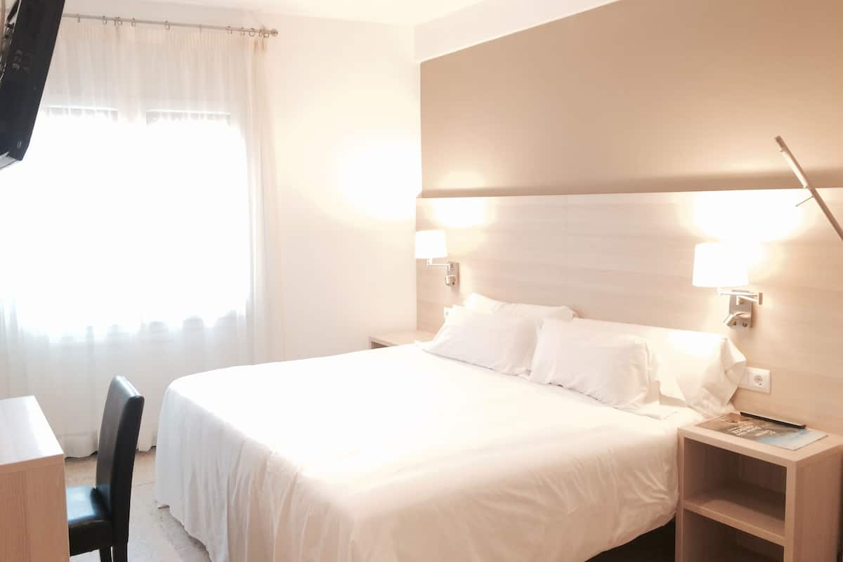 Quarto de hotel com uma cama grande, duas luminárias de cabeceira, uma televisão na parede, uma mesinha de cabeceira de madeira e uma cadeira.