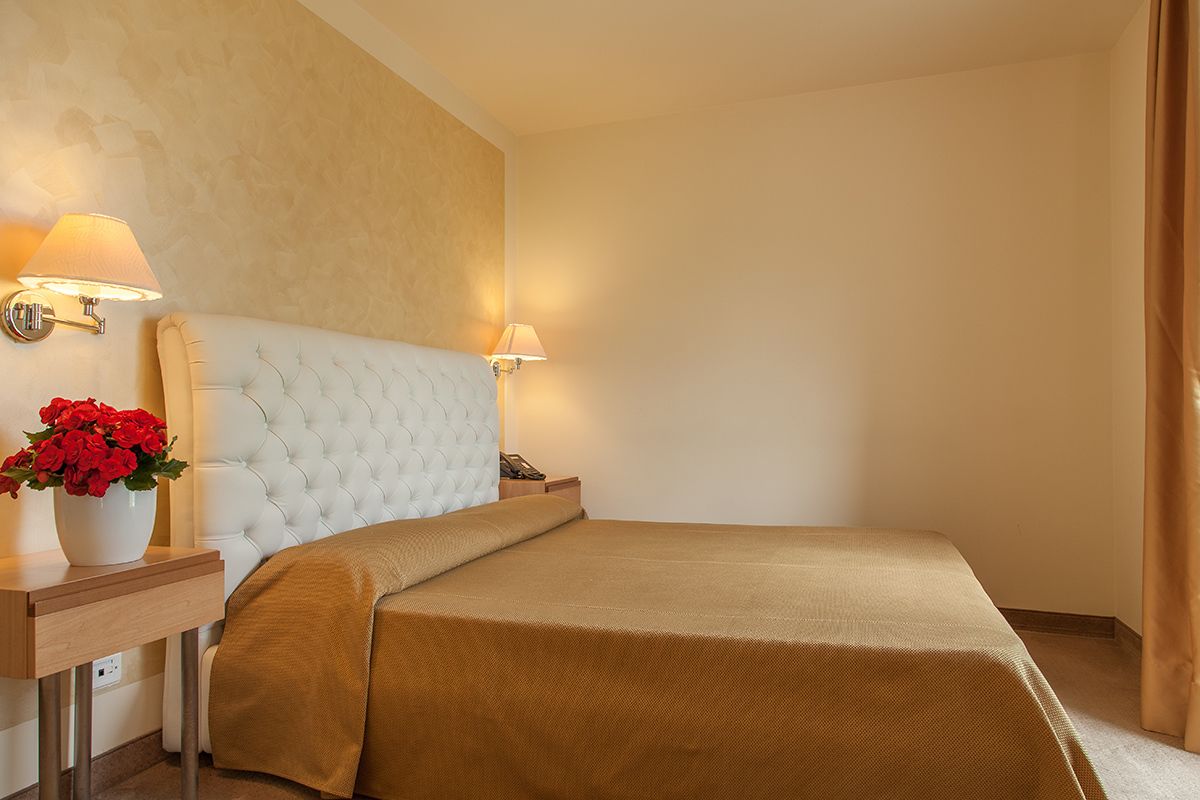 A hotel room with a tufted headboard, a beige bedspread, a bedside table with a lamp, and a vase of red flowers.