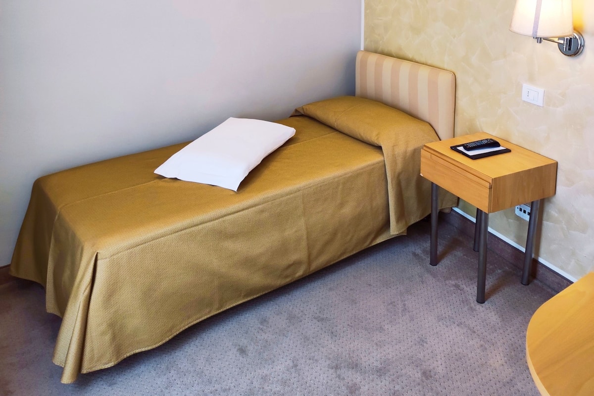 A small, neatly arranged hotel room with a single bed, a bedside table with a lamp, and a wall-mounted light.