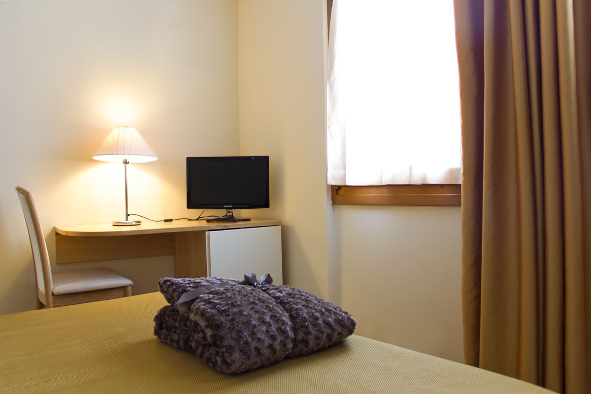 A hotel room with a bed, a desk with a computer, a lamp, and a window with curtains.