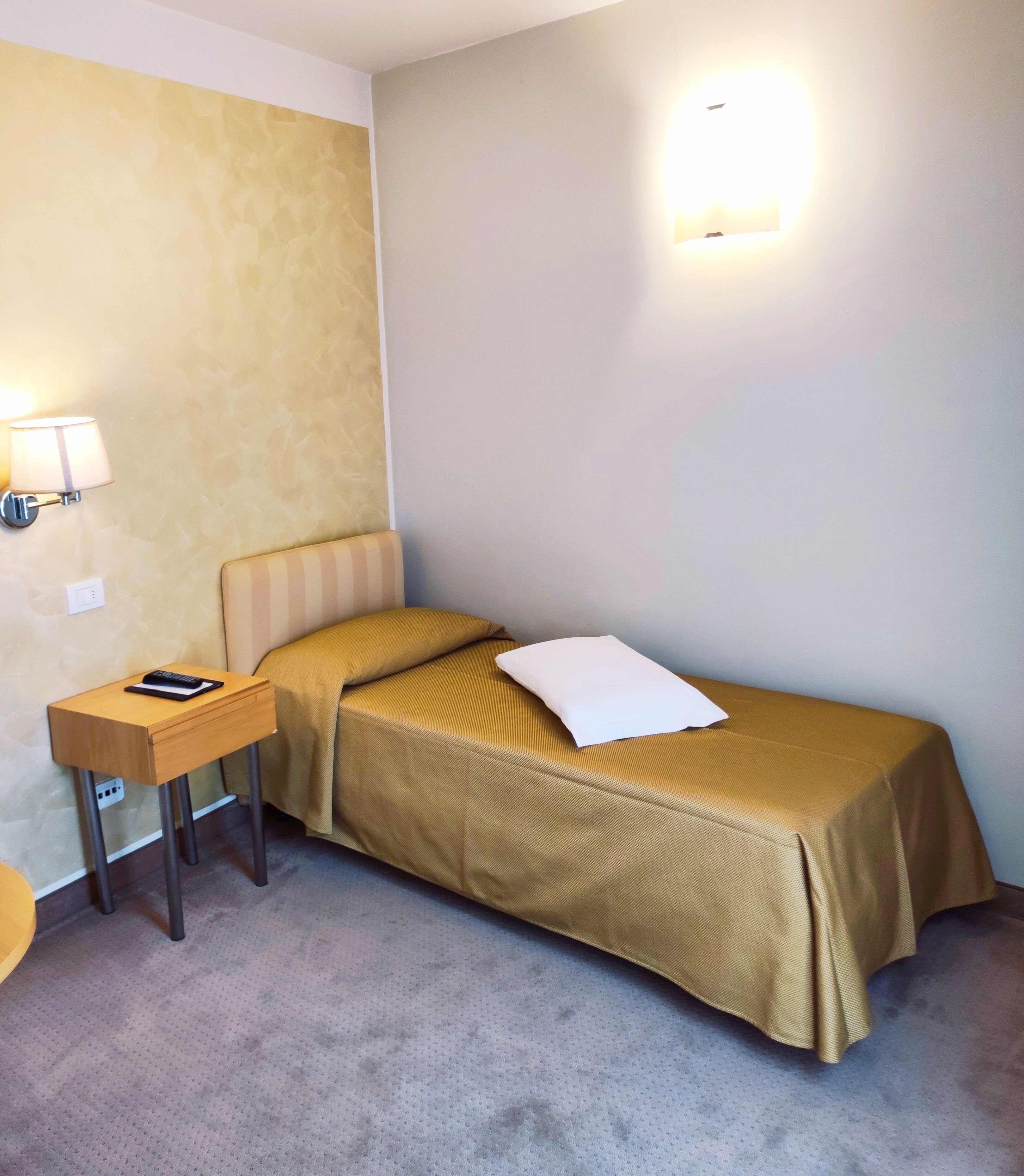 A small, single-bed room with a wooden nightstand, a lamp, and a wall-mounted light.