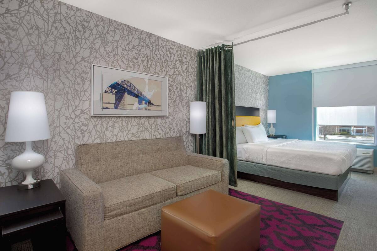 Habitación de hotel con sofá, cama, mesita de noche, lámpara, un cuadro en la pared y una ventana con cortinas.