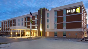 Un hotel moderno con un cartello ben visibile con la scritta "HOME 2", un ingresso coperto e una bandiera.