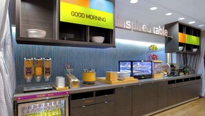 Una colazione a buffet con distributore per yogurt e latte, un cassetto per posate e un televisore che mostra un notiziario.