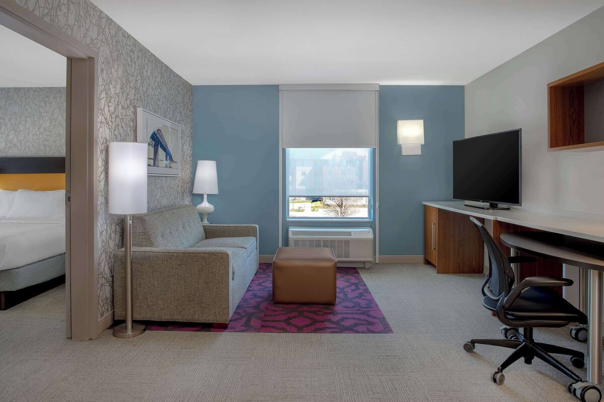 Habitación de hotel con cama, sofá, escritorio con televisor, ventana con vista y alfombra estampada.